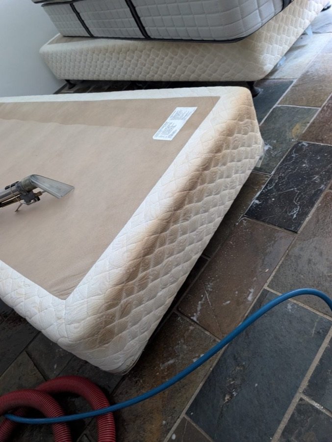 Before: white quilted mattress base with dark brown staining along its side, with a steam cleaning wand resting on top